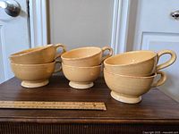 Six large beige ceramic handled bowls stacked on a wooden surface with a ruler for scale.