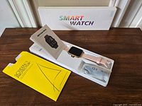Smart watch in open box with user manual and yellow envelope containing screen protectors on a wooden surface.