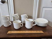 Photo showing 5 white ceramic ribbed tea cups stacked and one placed in front, 1 creamer pitcher, and 5 small plates stacked on right, ruler for scale