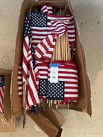 Box containing multiple American flag yard ornaments with wooden stakes in various sizes.