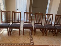 Front view of the six matching wooden chairs with brocade seats, arranged side by side on tile floor, showing overall appearance and condition.