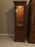 Full front view of the wooden display cabinet showing its tall, narrow design with upper glass door featuring grid pattern and light inside, below is a wooden door with similar grid pattern.