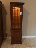 Full view of tall wooden display cabinet with upper glass door, decorative grid pattern, and lower wooden door with similar design, turned on interior light.