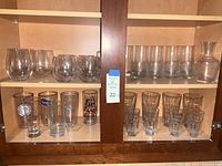 Clear glass barware assortment on wooden shelf with various glasses and a decanter.