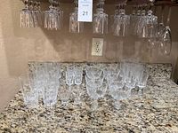 Photo of various crystal stemware and drinkware arranged on granite countertop with additional wine glasses hanging upside down above counter.