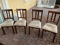 Four wooden dining chairs with beige upholstered seats positioned on outdoor brick floor. Chairs arranged side by side, backs facing camera.