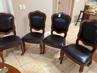 Four dark brown leather upholstered wooden dining chairs with nailhead trim, turned legs, two with casters, on tile floor near kitchen area.