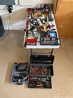 Overview of the hand tools assortment including socket sets, wrenches, screwdrivers, pliers, Dremel Multi Pro rotary tool, jumper cables, clamps, and levels arranged on a white folding table and on toolboxes below.