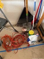 Photo showing step ladder, rake, shovel, extension cords, hummingbird feeder, and cleaning products on the floor of a garage.