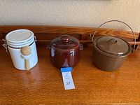 Photo of brown metal kettle, brown stoneware crock, and white ceramic canister with wooden spoon placed on wooden surface against wall.