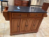 Front angled view showing the kitchen island cart's wood finish, granite top, two front drawers open, and four cabinet doors below.