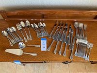 Full set of Oneida Deluxe stainless flatware arranged on wooden surface showing all forks, knives, spoons, and serving utensils.