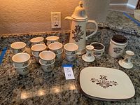 Wide view of lot showing Pfaltzgraff Village teapot, custard cups, candle holders, buffet plates, and coffee/sweetener set on kitchen counter.