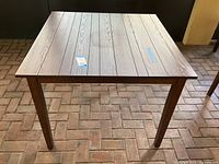 Square outdoor wood table from a top-front angle on brick flooring showing wood plank top and four legs