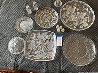 Top-down image of six etched glass platters and plates, plus three pairs of salt and pepper shakers with metal tops on a textured blanket.