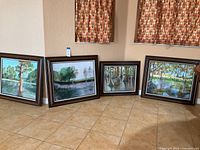 Wide shot showing all four framed paintings leaning against wall under window with patterned curtains, tiled floor beneath