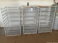 Three white metal wire storage units in a garage setting, each with multiple pull-out wire drawers. One unit from left to right is missing the top panel. Units are used and show visible dirt.