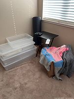 Stack of three large clear plastic storage containers with lids, small black folding table, essential oil kit on table, and two hoodies on a box beside them.