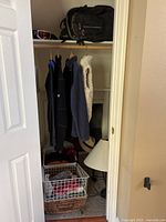 Closet view showing hanging clothing items, black backpack on shelf, pink ski goggles, white table lamp on floor, and storage basket with textiles in closet floor.