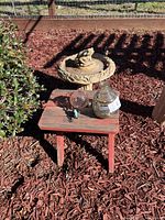 Photo showing concrete birdbath with frog design, red wooden side table, two glass bird feeders on side table, placed outdoors on mulch.