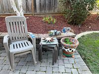 Two beige plastic outdoor chairs with three brown plastic side tables stacked between them, colorful flower pot on a stand, metal garden decor and animal figurines on the ground