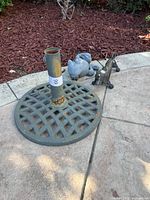 Photo showing round lattice-pattern cast iron umbrella stand with a vertical rusted tube holder, placed on concrete.