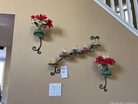 Three wall-mounted metal sconces shown on a beige wall. Two have green glass holders with faux flowers, one holds six candles on circular holders.