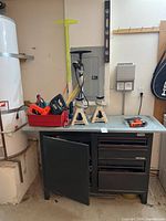 Wide photo showing entire metal workbench with drawers and cabinet, red tool tray on top with power tools, and three jack stands on the bench surface.