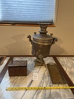 Photo showing samovar, lock box, and brass tray with measurement tape for scale on table.
