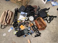 Wide view of assorted vintage and modern camera equipment, including fabric and leather camera bags, a tripod, camera batteries, and accessories scattered on a floor.