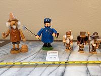 Seven wooden figurines lined up including four small nutcrackers and three larger smokers on a marble surface with tape measure showing size approx. 4-7 inches tall.