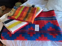 Three assorted wool blankets and throws displayed on a bed showing red and blue Navajo design, white Faribo striped throw, and red Curlew Weavers throw with fringes.