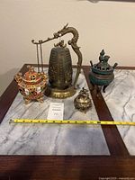 Four vintage Chinese items on table: brass dragon bell, bronze incense burner with bird lid, porcelain Satsuma incense burner, and smaller brass incense burner with measuring tape showing lengths.