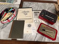 Overview photo showing calligraphy dip pens in a red tin box, boxed calligraphy pen set, instructional booklets on calligraphy paper and writing nibs, and a brown notebook.