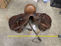 Top-down view of the brown Crosby English saddle showing stirrups and a riding crop alongside a tape measure for size reference.