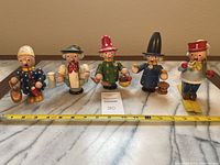 Five wooden smoker figurines arranged in a row, showing their full front view with measuring tape indicating size, about 5-6 inches tall.