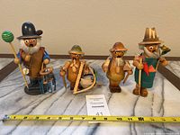 Five wooden smokers figurines displayed in a row with a measuring tape showing sizes ranging approximately from 5 to 9 inches tall.