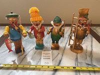 Four wooden smoker figurines lined up on marble surface with measuring tape showing heights and lot tag