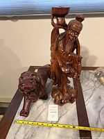 Photo showing carved wooden lion figure and Shou Lao figure side by side on table with measuring tape for scale.