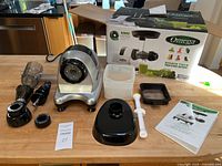 Omega MM900HDS juicer and all main components laid out on a wooden table with original box in the background, shows motor base, containers, manual, and various attachments.