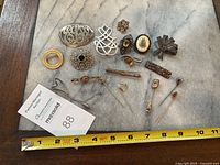 Top-down view of the assortment of vintage stick pins, brooch components, and brooches on a marble surface with a measuring tape indicating sizes up to 10 inches.