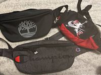 Photo showing three belt bags: Timberland black with silver tree logo, Nike Air black and red with drawstring top, and grey Champion with embroidered logo.