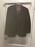 Front view of the Calvin Klein men's wool suit jacket hanging on a hanger against white blinds. Dark color, single-breasted with flap pockets.