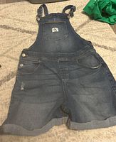 Full front view of dark blue denim Calvin Klein overalls laid flat showing adjustable straps, bib pocket with CK logo, side button closures, length to mid-thigh with rolled hems.