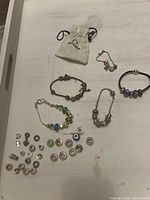 View of three charm bracelets with assorted loose charms spread out on surface, including a small Pandora pouch.