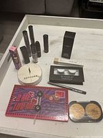 Makeup items arranged on a table including eyeshadow palette, face powder, false eyelashes, lipsticks, and makeup tubes with cosmetic bag visible.