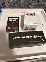 Three adult card games: 'What Do You Meme?' original white boxed game, 'What Do You Meme?' NSFW Expansion Deck in black, and 'Cards Against Disney' in a black box.