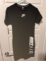 Full front view of black Nike Air dress hanging on hanger with door background. Shows short sleeves, crew neck and white Nike Air logos on chest and lower side.