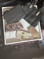 Five designer bags and wallets displayed in a white tray: two black handbags, one tan wallet, one snake-skin patterned clutch, and a cream clutch.