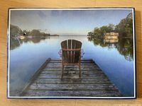 Framed photographic artwork showing wooden dock with single Muskoka chair facing calm lake surrounded by trees and boathouse.
